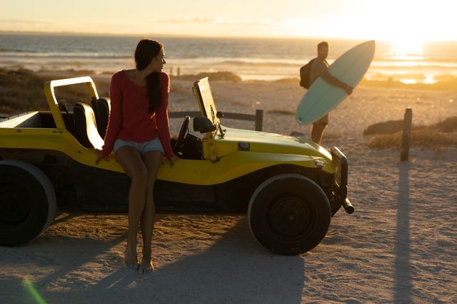 Woman sitting on yellow beach buggy, watching man carrying surfboard towards sea at sunset. Perfect for travel blogs, summer vacation promotions, romantic getaway advertisements, and adventure lifestyle content.