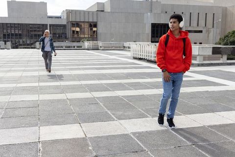 Teen in red hoodie listening to music while walking across urban concrete campus plaza