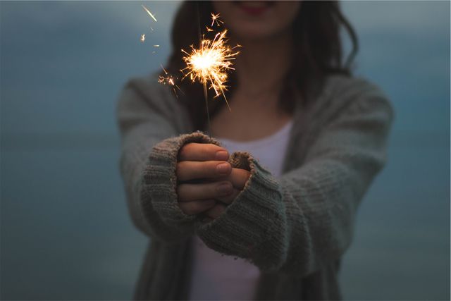 This image shows a young woman wearing casual clothing holding a sparkler outdoors, creating a warm and festive atmosphere. This can be used for events promotions, holiday-themed designs, inspiration posts, and social media campaigns emphasizing celebration and joy.