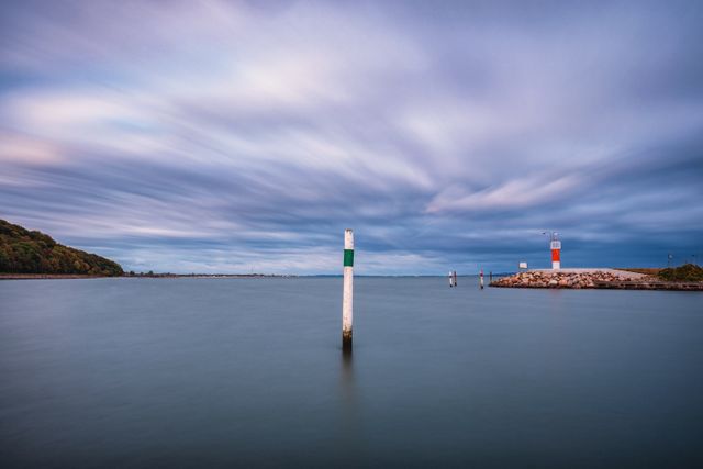 This image features a calm coastal harbor during sunrise with wooden pylons serving as maritime navigation markers. The serene water and the soft morning light cast an ethereal glow over the scene. This peaceful coastal landscape with its quiet waters and cloudy sky makes it perfect for projects related to relaxation, travel inspiration, or coastal living.