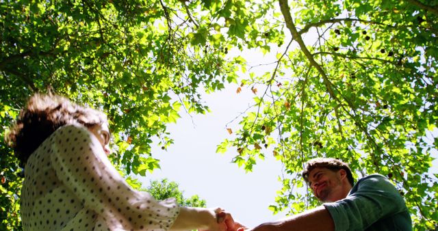 A young Caucasian man and woman are holding hands and spinning around joyfully under a canopy of green leaves, with copy space. Their playful interaction captures a moment of carefree happiness and connection in a natural setting.