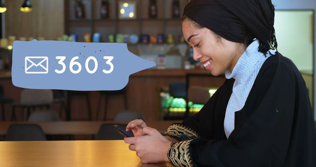 Happy woman in traditional attire checking her smartphone while sitting in a coffee shop, displaying a message notification indicating 3603 messages. Useful for themes related to social media, communication, technology, and modern lifestyle in coffee shop settings.