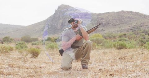 Kneeling hunter scanning arid scrubland with binoculars and shotgun, wearing khaki pants and cap