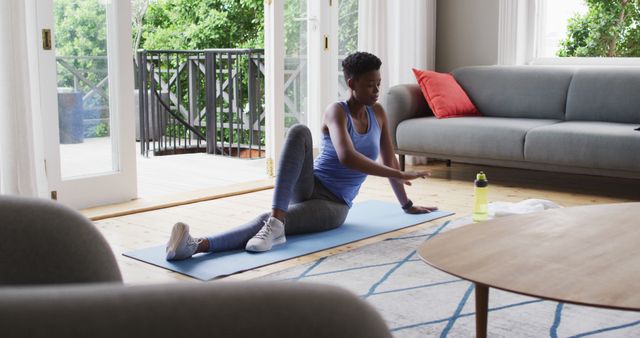 Image shows a young woman in athletic wear doing a stretching routine on a yoga mat in her living room. The room is bright with natural light pouring in through large windows, enhancing the homely and serene atmosphere. Ideal for use in content about home workouts, fitness and wellness routines, personal training, and indoor health activities.