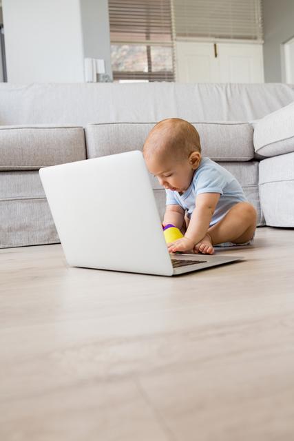 Baby boy sitting on floor in living room, playing with laptop. Ideal for use in articles about childhood development, technology in early education, family life, and modern parenting. Suitable for blogs, websites, and advertisements focusing on children, technology, and home environments.