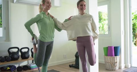 Two women stretching in sunlit home gym wearing leggings and activewear fitness lifestyle