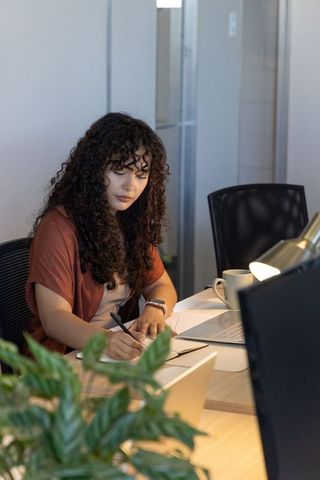 Young woman writing in notebook while working late at office desk with laptop and lamp