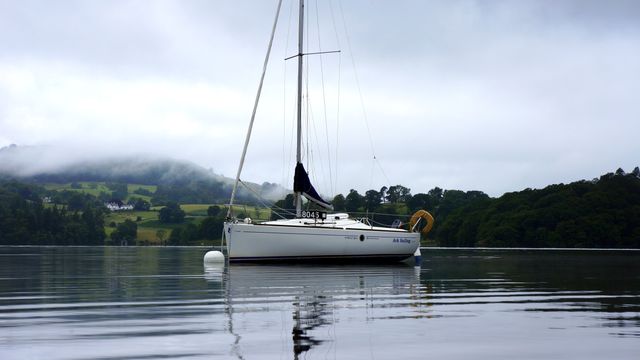 Sailboat anchored on serene lake with tranquil water reflecting the surroundings. Hilly landscape shrouded in mist adds a scenic and peaceful backdrop, ideal for promoting travel, tourism, or outdoor adventures.