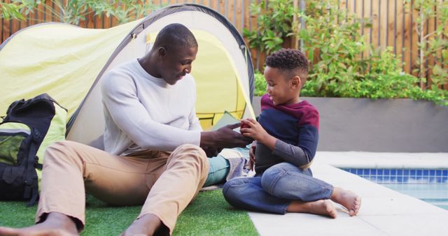 Father and son spending quality time outdoors, sitting next to a tent on grass near a pool. The father in a white sweater and beige pants looks lovingly at his son, who is handing him a piece of paper. Green plants in the background lend a serene atmosphere. This could be used for family-oriented advertisements, or articles on parenting and outdoor activities.