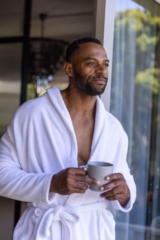 Man standing in white bathrobe holding morning coffee at glass doorway, contemplating view