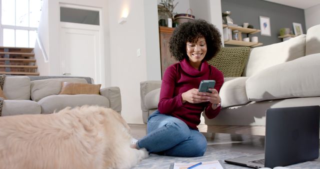 Young African American woman sitting on floor in modern living room, using smartphone and smiling while a dog lies beside her. Laptop and papers scattered, emphasizing relaxed and casual setting. Perfect for themes around home life, remote work, technology use, and pet companionship.