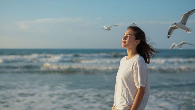 Young woman standing on sunlit beach facing ocean breeze with seagulls and calm expression