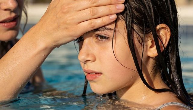 Caregiver checking child forehead in pool closeup, sunlit water safety heat concern