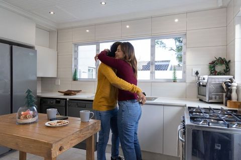 Two women hugging in bright modern kitchen sharing cozy moment with coffee and cookies