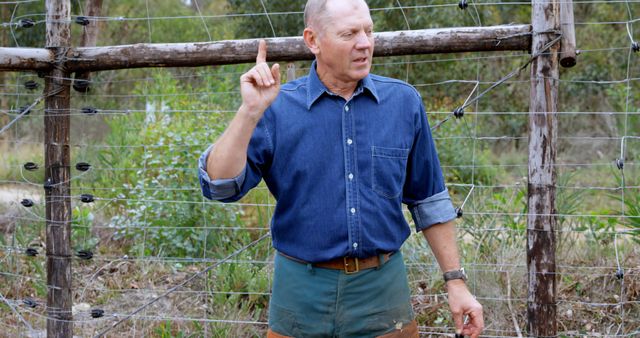 Farmer standing outdoors near wire fence, explaining sustainable farming techniques by pointing with index finger. Suitable for agricultural education, rural lifestyle promotions, environmental sustainability campaigns, and farming training materials.