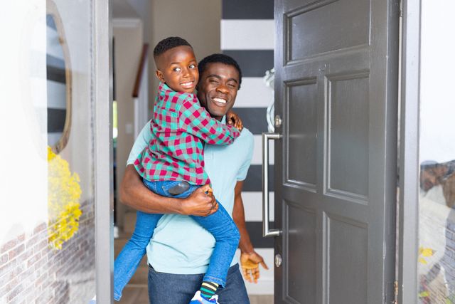 Happy african american father holding son at open front door of home, smiling, copy space. Family, togetherness, domestic life and happiness concept.