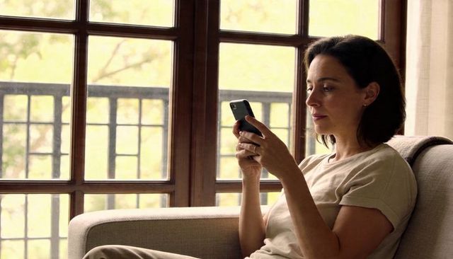 Woman using smartphone while relaxing on couch by sunlit wooden window