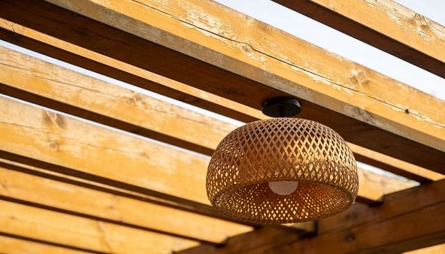 Woven rattan pendant lamp casting warm linear shadows under sunlit wooden pergola