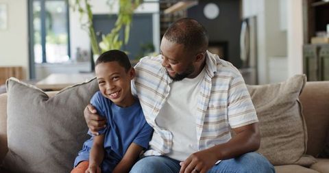 African american father sharing warm laugh with son on couch, cozy family bonding moment