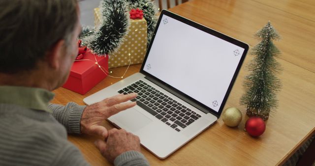 Senior man using a laptop at a wooden table decorated with Christmas ornaments and wrapped gifts. Ideal for holiday-themed technology usage, online celebrations, virtual gatherings, or remote family connections during holidays.