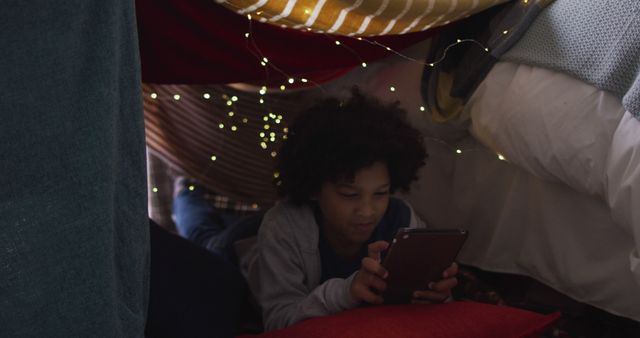Young boy engaging with tablet in a cozy DIY blanket fort illuminated by soft string lights. Perfect for themes of childhood, leisure, technology, indoor activities, and cozy settings.