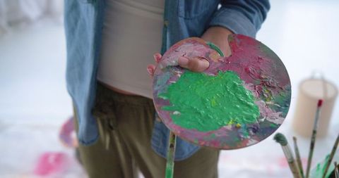 Mature woman holding paint palette and brush wearing denim shirt in bright art studio