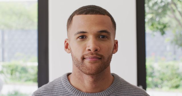 This image shows a confident young man posing indoors while looking directly at the camera. He is wearing a casual gray sweater, adding a relaxed and approachable vibe to the scene. The neutral, modern background with large windows and greenery outdoors can make this image ideal for various uses, including advertising, lifestyle blogs, and corporate media. Its straightforward and professional appearance will suit materials wanting to convey trust, modernity, and relatability.