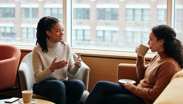 Two women chatting over coffee in cozy lounge by window, friendly conversation