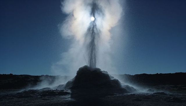 Erupting geyser casting haloed sun through rising steam plume over rocky geothermal basin