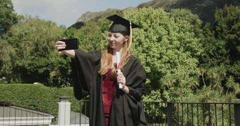 Graduate taking selfie on balcony wearing cap and gown holding diploma