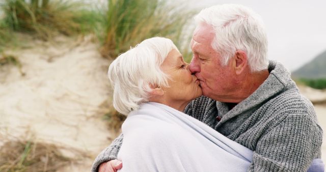 A senior Caucasian couple shares a tender kiss on a beach, wrapped in a cozy blanket, with copy space. Their affectionate moment captures a timeless love and companionship.