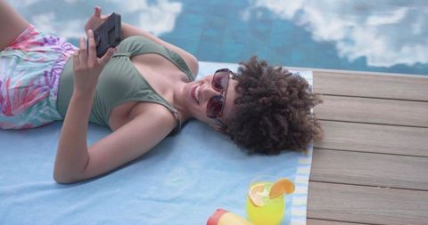 Reclining african american woman smiling and scrolling phone poolside with tropical drink