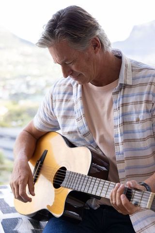 Mature man playing acoustic guitar on sunlit balcony overlooking hills, casual musician