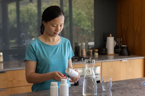Asian woman preparing daily medication on kitchen island with water and pill bottles