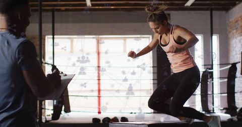 Female athlete performing explosive plyometric box jump inside boxing ring with coach