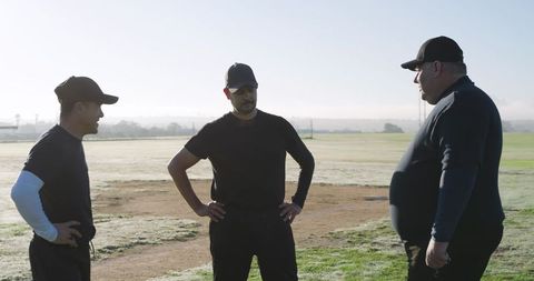 Mature baseball teammates discussing strategy on dew-covered grass field at dawn