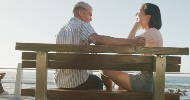 Couple sitting closely on wooden bench by ocean, enjoying time together under sunlight. Use for themes like relationships, outdoor relaxation, vacation, and beach settings.