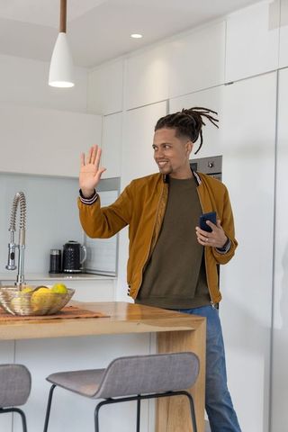 African american man waving while holding smartphone in modern kitchen with bomber jacket