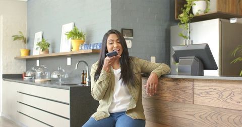 Indian woman speaking to smartphone while ordering at cafe counter leaning on wooden bar POS