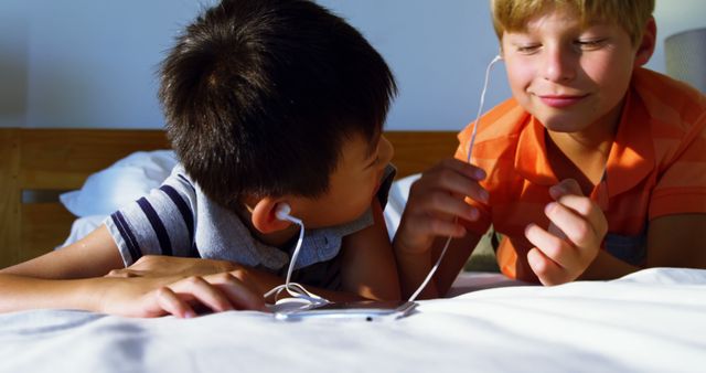 Boys enjoying music together and smiling while laying on a bed, sharing earphones. Perfect for illustrating concepts of friendship, childhood, and technology use among youth. Ideal for articles, blogs, or advertisements promoting children’s products, friendship, and play.