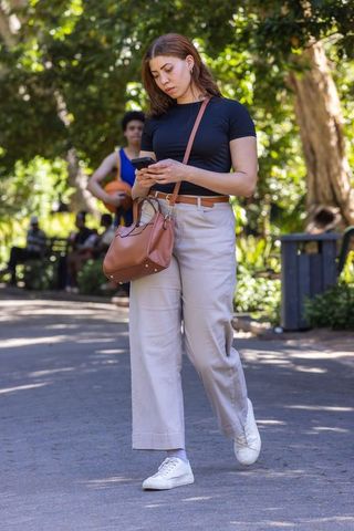 Young woman walking through park checking smartphone while carrying crossbody bag