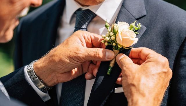 Groom boutonniere fastening with pale peach rose on navy suit lapel close-up
