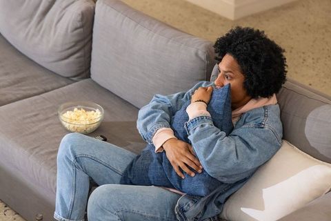 African american man hugging pillow on sofa watching tv with bowl of popcorn