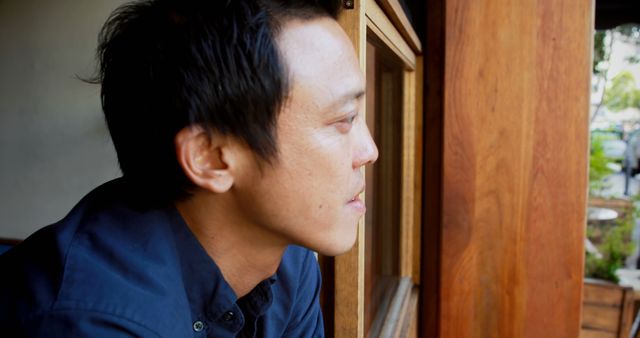 An Asian man in his middle age looks thoughtfully out of a window, creating a serene moment perfect for promoting themes of introspection or decision making. His expression of quiet contemplation can be used in healthcare, mental health, or business contexts, such as for advertisements discussing personal journeys, future plans, or waiting for important events. The copy space offers room for overlays or messages related to personal projects or reflective storytelling.