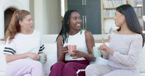 Three women chatting and sipping mugs on cozy living room sofa, casual conversation