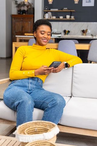 African american woman relaxing on sofa scrolling smartphone in open-plan lounge