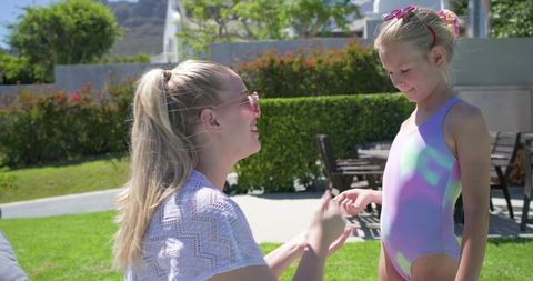 Mother checking daughter's swimsuit and sunglasses on sunlit backyard lawn, family bonding