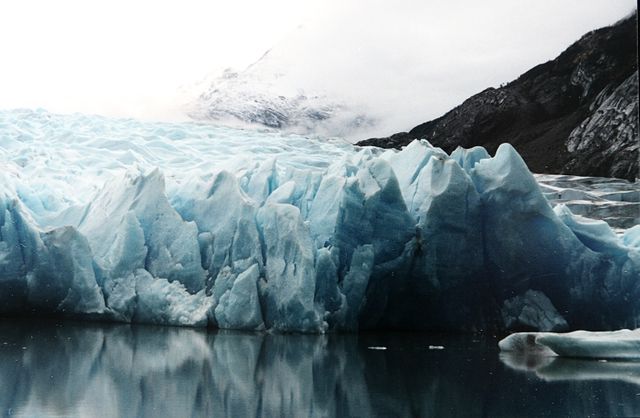 The image portrays a magnificent glacier with striking blue ice, reflecting in calm waters. The mountainous terrain adds to the awe-inspiring natural scenery, making this image suitable for projects related to nature, climate change, and travel. This serene and majestic landscape can also be used in environmental campaigns highlighting glacial retreat and global warming.
