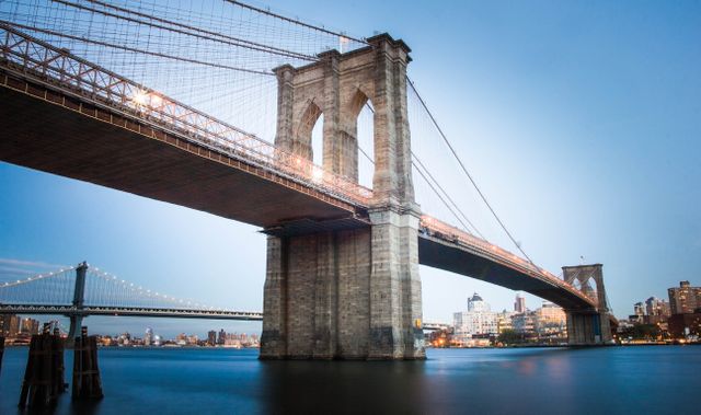 Brooklyn Bridge spanning over calm East River during dusk in New York City with city lights in background. Suitable for travel promotions, urban planning presentations, tourism brochures, and architectural showcases.