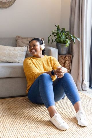 Smiling african american woman sitting on rug wearing mustard sweater and headphones